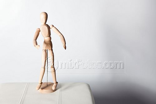 An articulated wooden mannequin stands on a round base at the edge of a white surface next to a light-coloured wall.