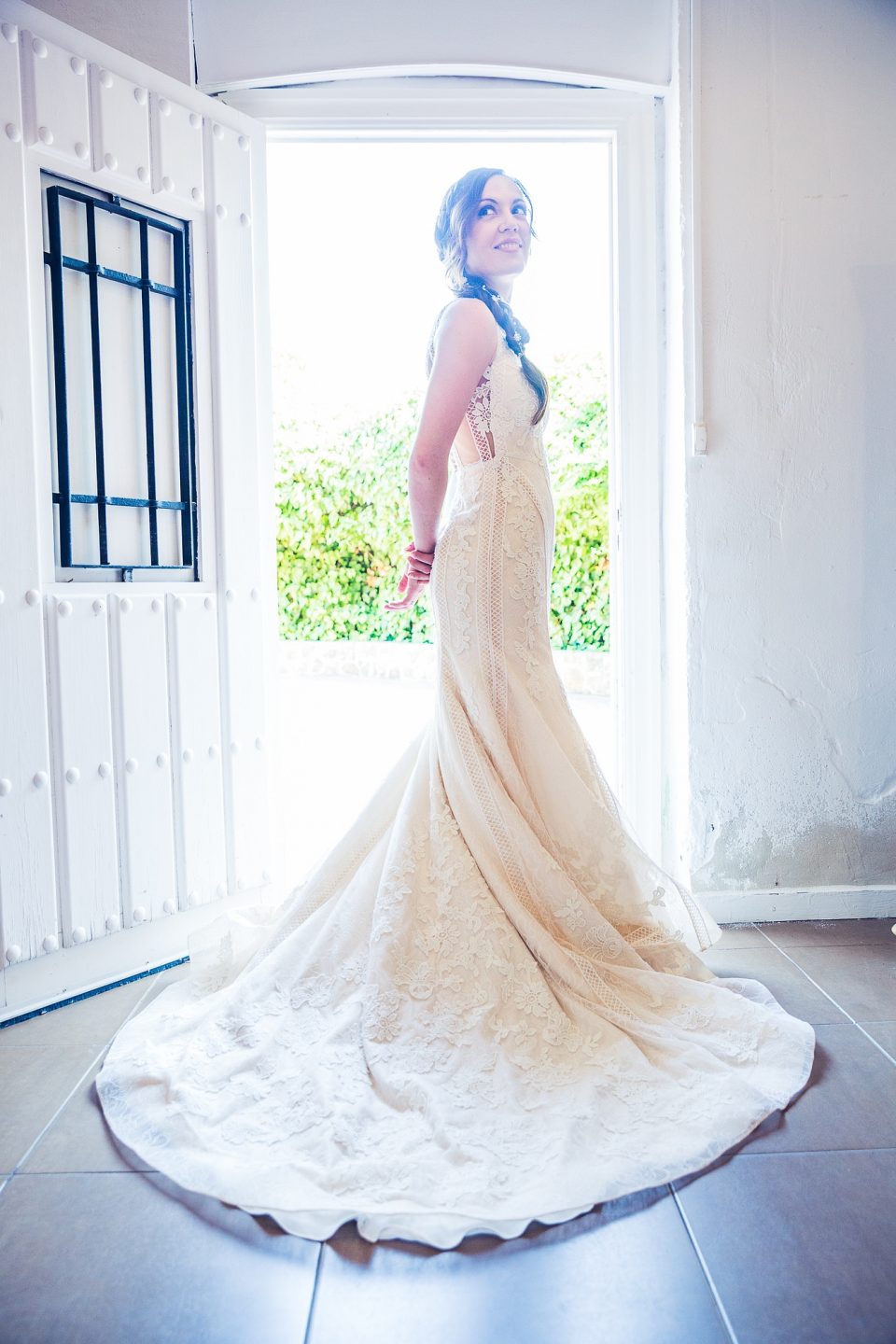 Mujer con vestido de novia largo de encaje y cola amplia, de pie junto a una puerta abierta en el interior de una casa.