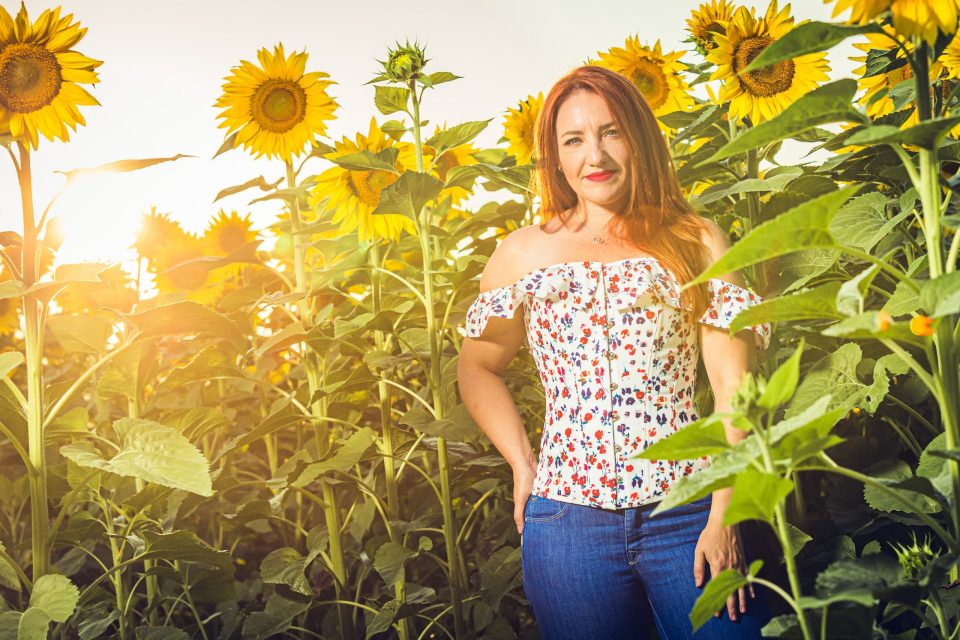 Woman standing among tall, lush sunflowers, wearing a white floral-print blouse and blue jeans.