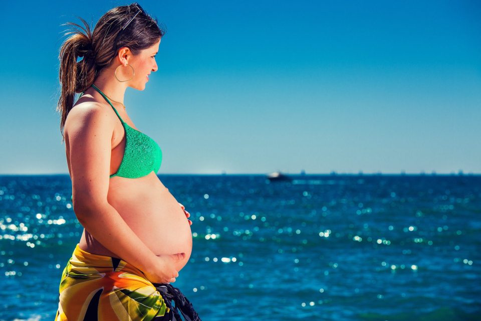 Mujer embarazada en perfil, junto al mar, con bikini verde y pareo colorido, mientras sostiene suavemente su vientre.