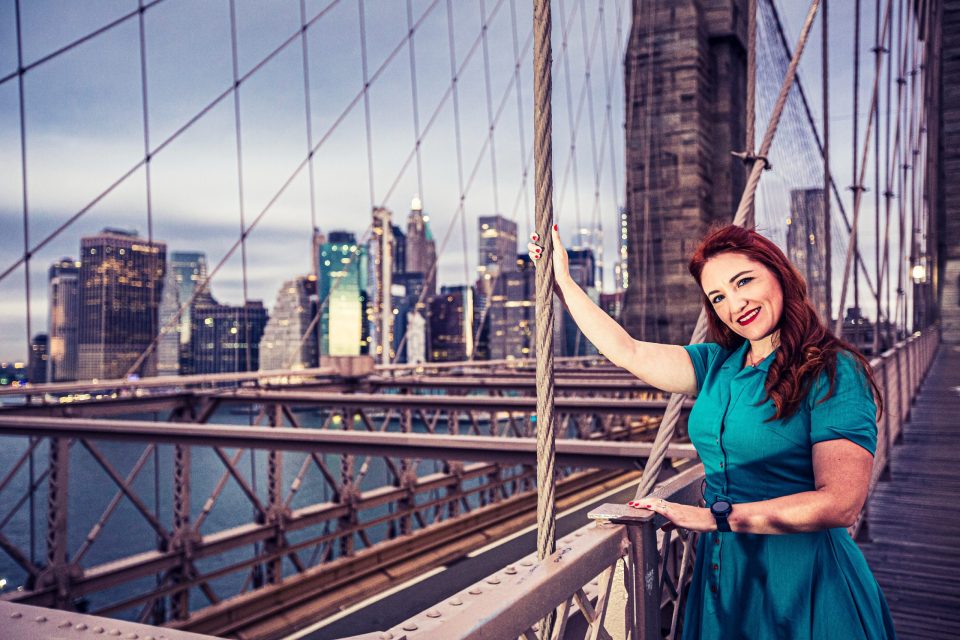 A smiling woman in a turquoise dress poses beside the cables and railing of a suspension bridge at sunset.