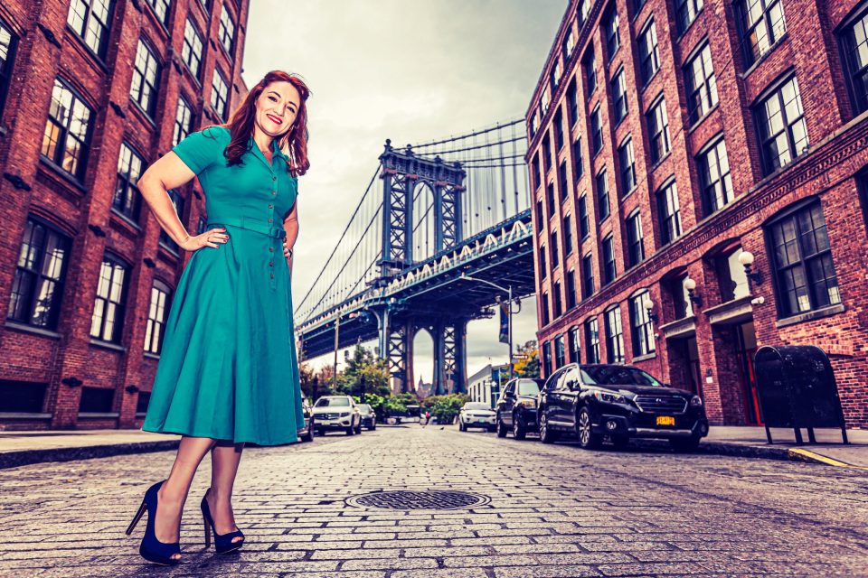 A smiling woman in a turquoise dress and high heels poses on a cobbled street between brick buildings.