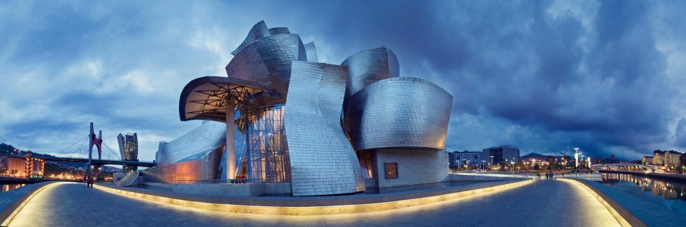 The Guggenheim Museum Bilbao is shown in a panoramic photograph, with a contemporary building featuring curved volumes clad in panels.