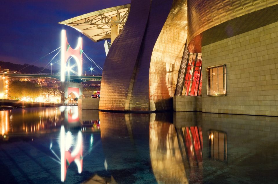 The Guggenheim Museum Bilbao is shown at night, with its contemporary building featuring curved forms and metal cladding.