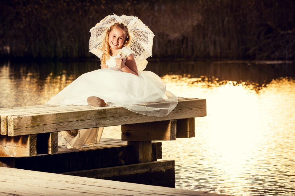 Niña con vestido blanco de comunión y sombrilla de encaje, sentada sobre un embarcadero de madera junto a un lago.