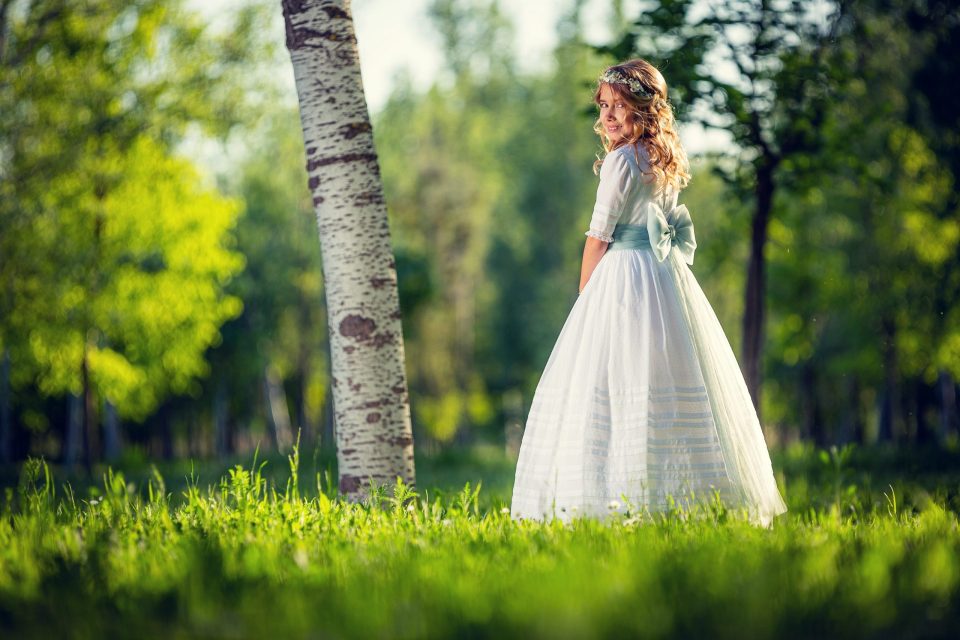 Niña con vestido blanco largo y cinturón verde, adornada con una corona floral, de pie entre la hierba de un parque arbolado.