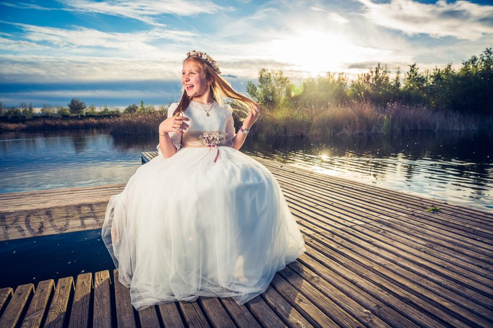 Niña con vestido blanco y corona floral sentada sobre un embarcadero de madera junto a un estanque.
