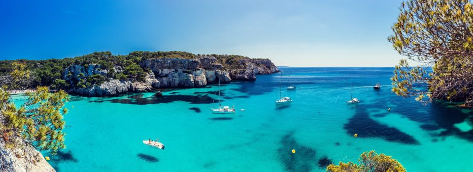 A panoramic landscape shows a cove with turquoise waters, surrounded by rocky cliffs covered in Mediterranean vegetation.