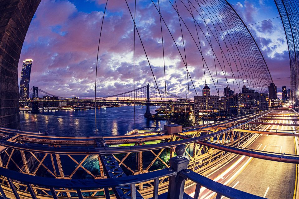A night-time cityscape viewed from a suspension bridge, with a dense network of cables and metal structures in the foreground.