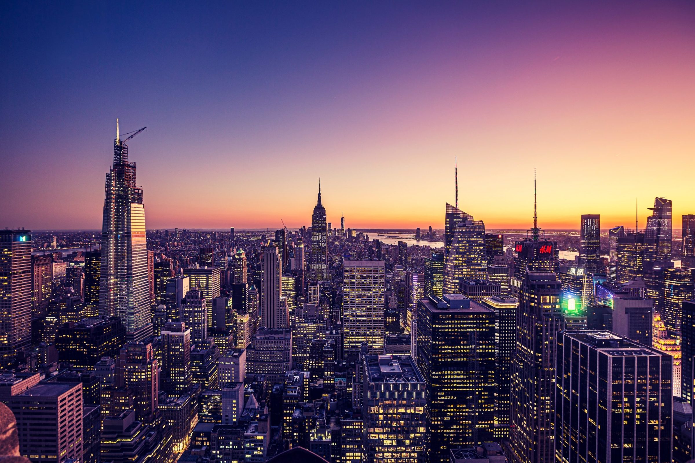 A panoramic urban landscape at sunset shows numerous illuminated skyscrapers.