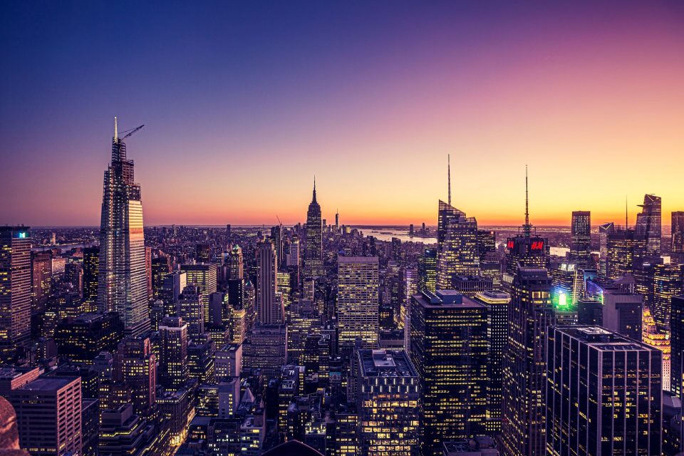 A panoramic urban landscape at sunset shows numerous illuminated skyscrapers.