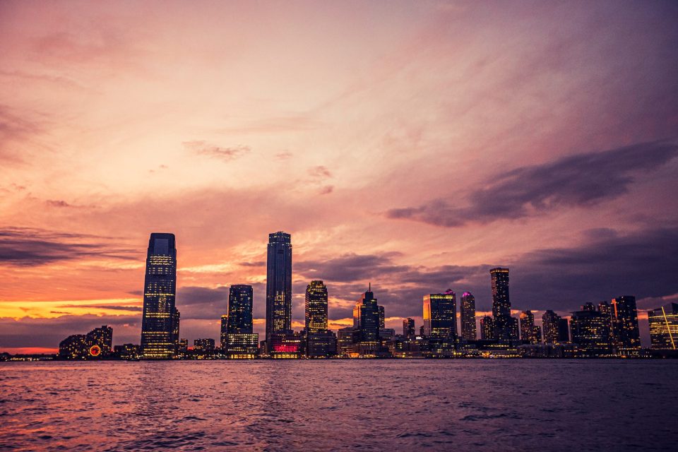 A panoramic view shows a city skyline by the water at sunset.