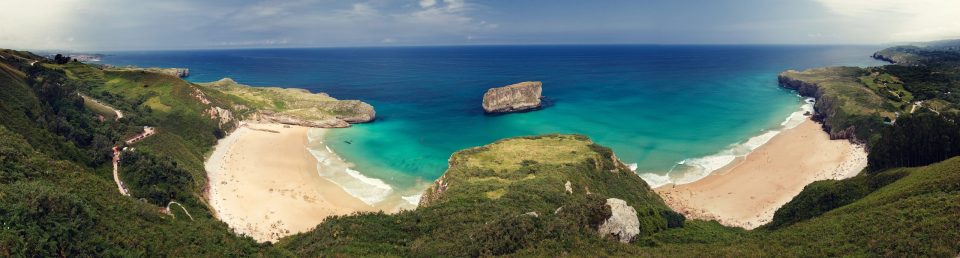 Panorama of a coastline with two coves of pale sand, green cliffs and blue and turquoise waters.