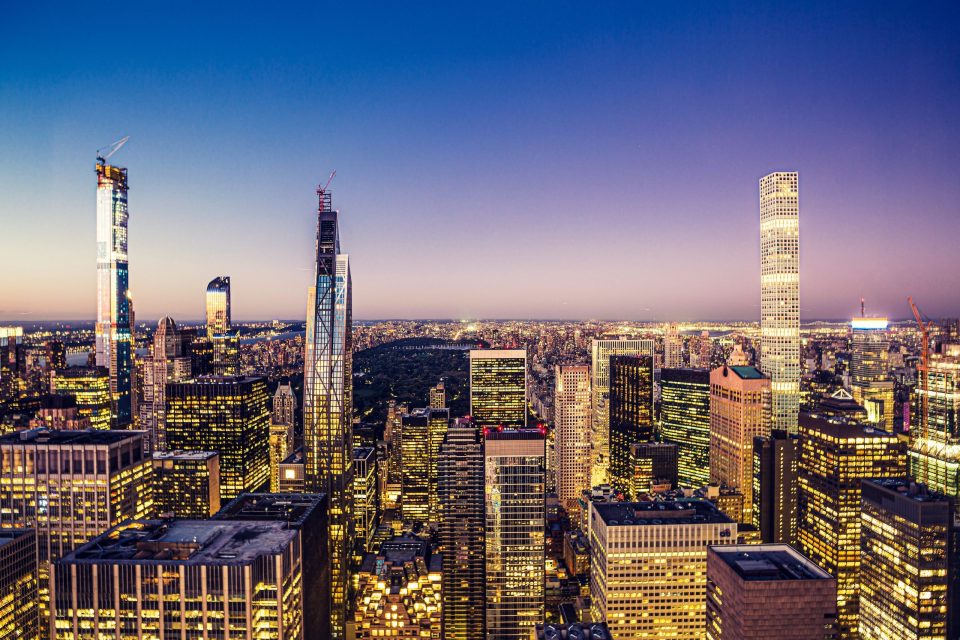 An urban panorama at dusk with numerous illuminated skyscrapers.
