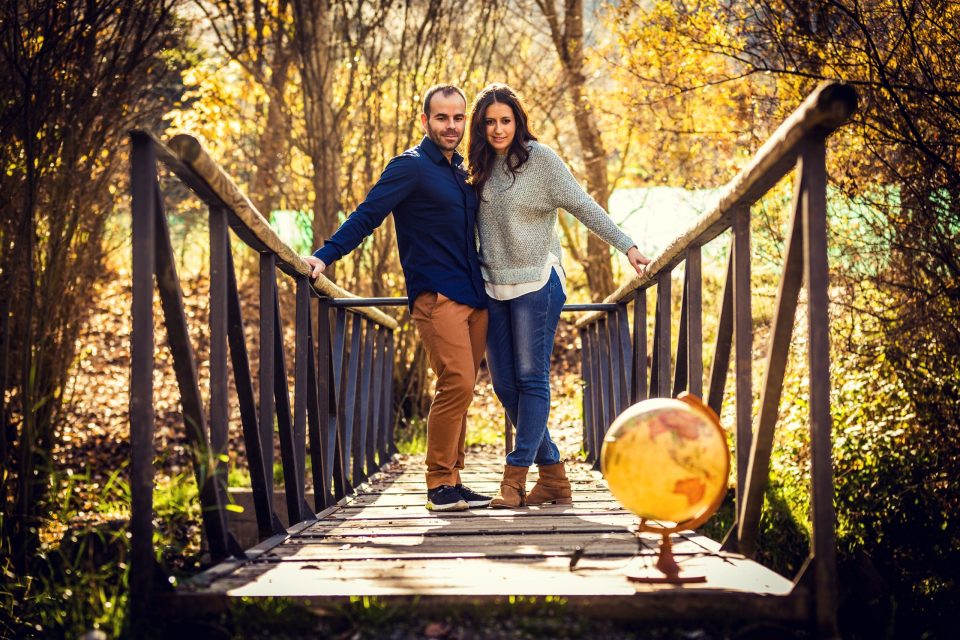 A couple walks along a wooden footbridge in a woodland setting with autumn leaves and warm light.