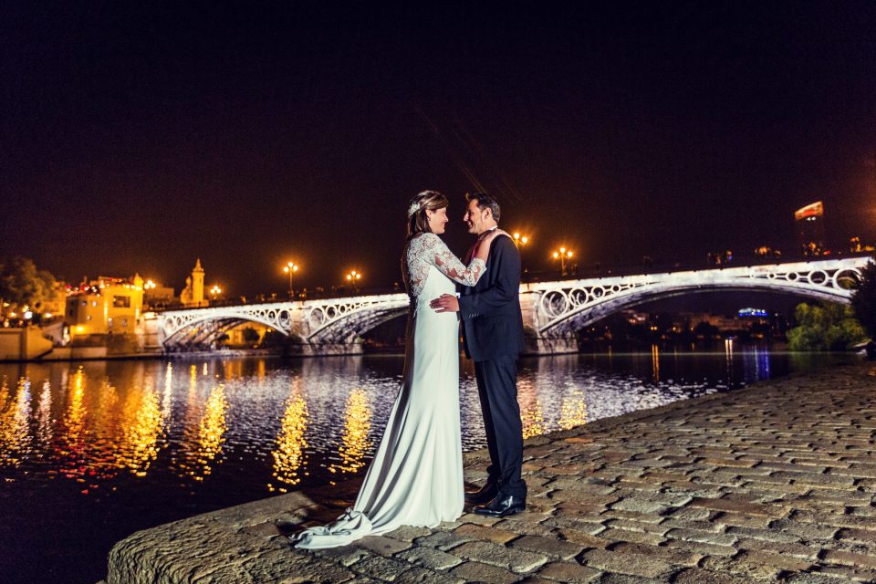 A couple in wedding attire embraces beside a river at night.