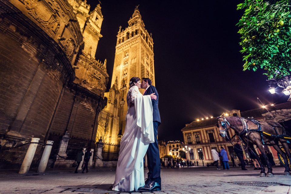 A couple in wedding attire embrace in front of an ornate, illuminated historic building at night.