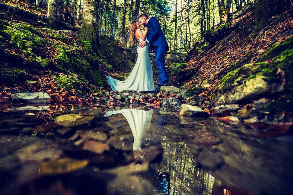 Pareja con vestimenta nupcial se besa en un arroyo de un bosque, rodeada de rocas, musgo, hojas y árboles altos.