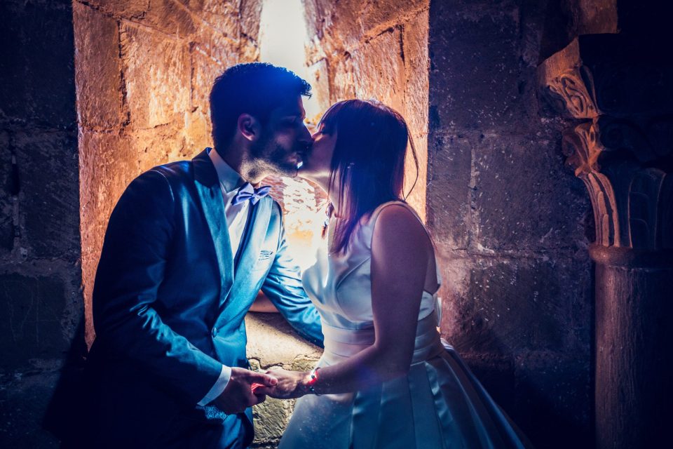 A couple in wedding attire kiss while holding hands in front of a lit window.