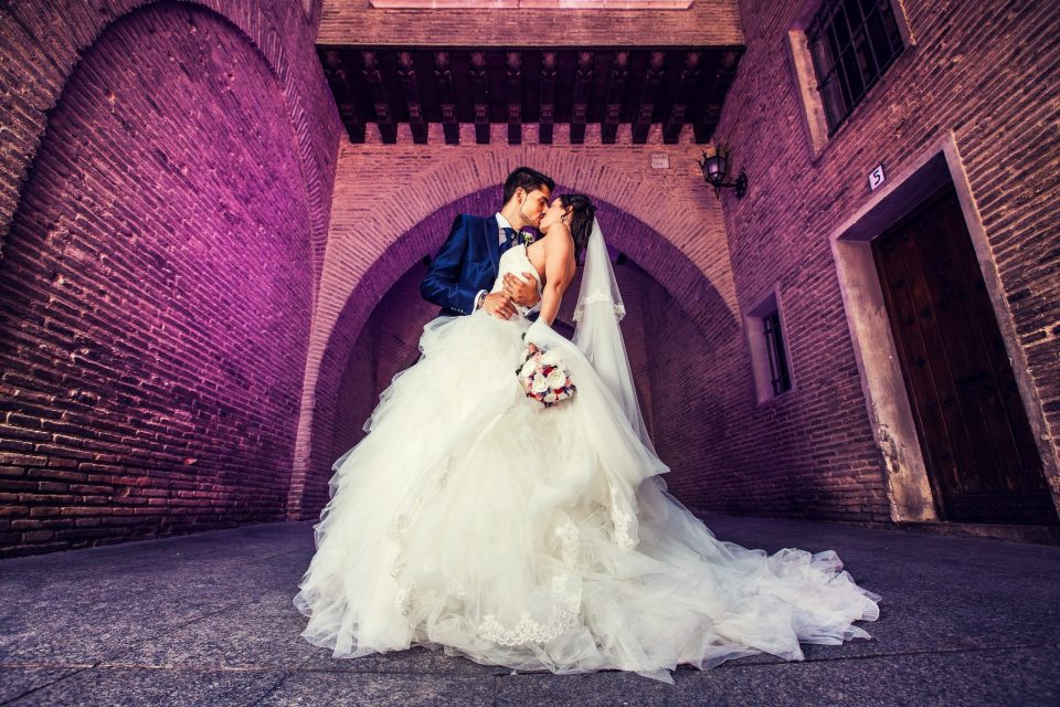 Pareja de novios besándose bajo un gran arco de ladrillo.
