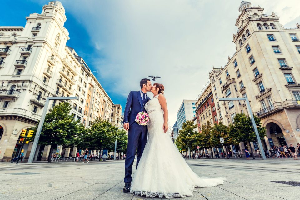Pareja de novios besándose en el centro de una plaza urbana, rodeada de edificios históricos, árboles.