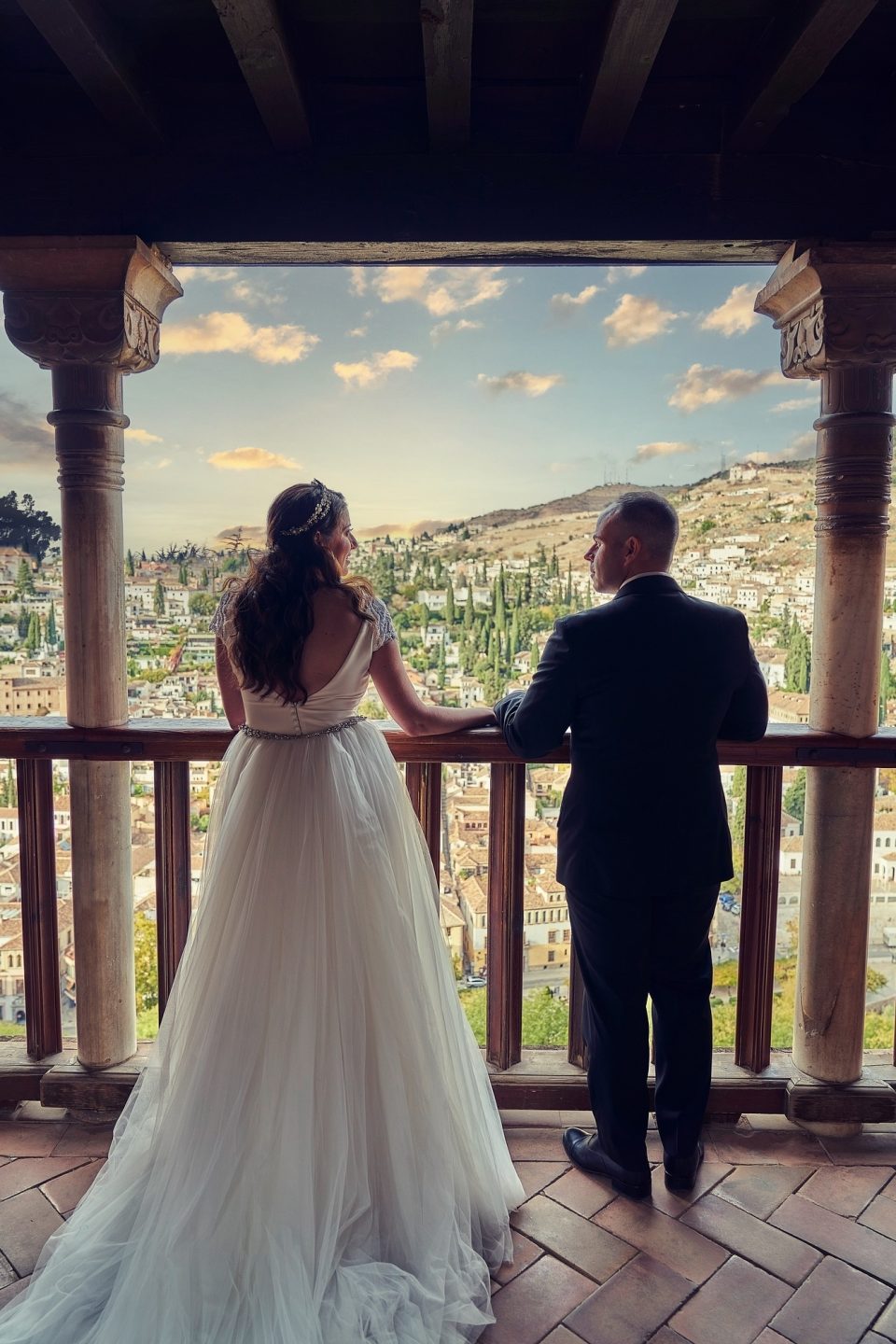 Pareja de novios contemplando una ciudad asentada en una ladera desde un mirador de piedra con columnas ornamentadas.