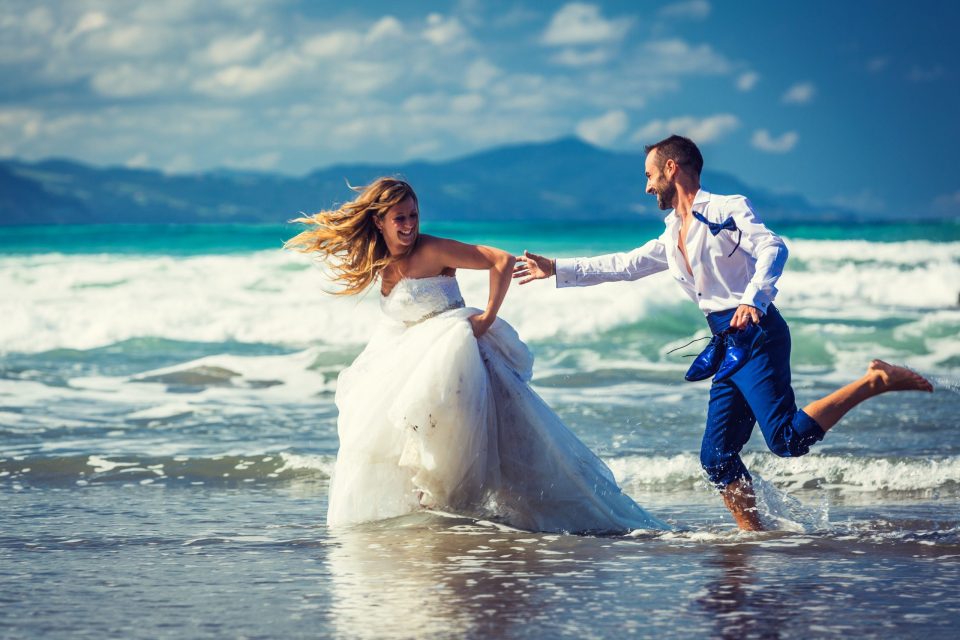 Pareja de novios corriendo y tomándose de la mano dentro del agua poco profunda de una playa.