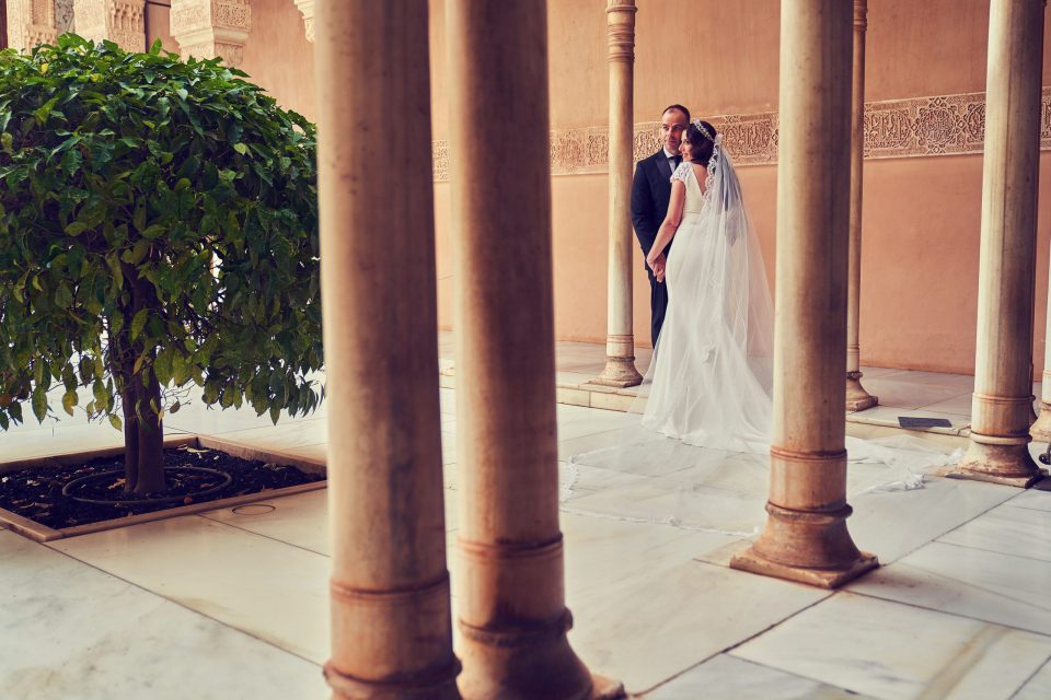 Pareja de novios en un patio monumental con columnas, arcos decorados y paredes ornamentadas.
