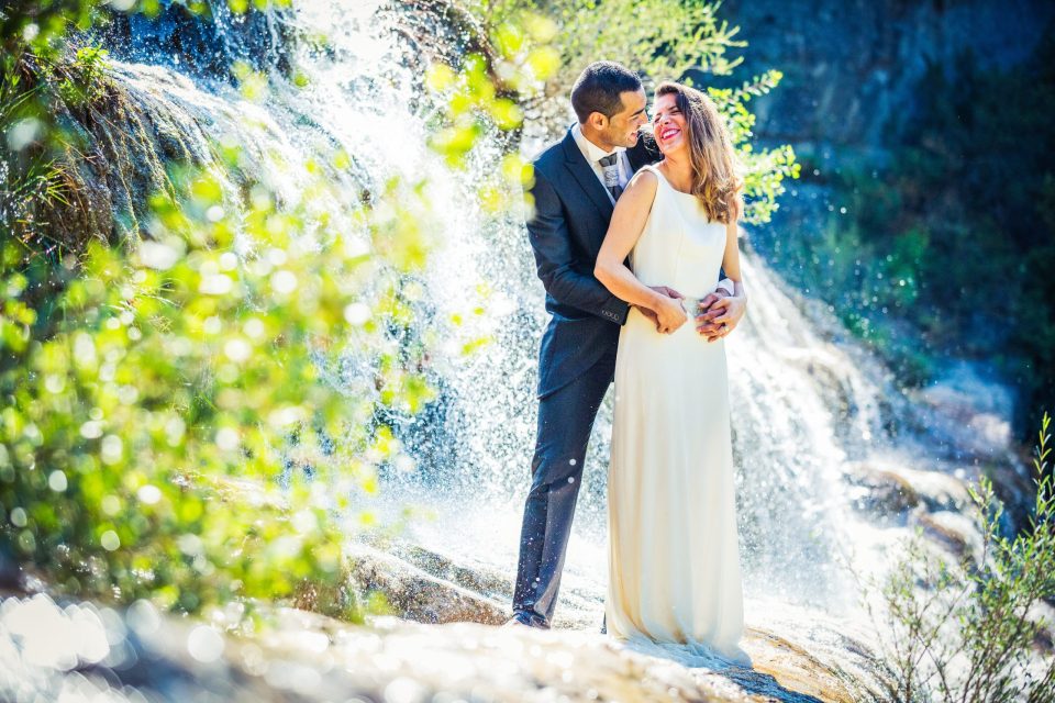 Pareja de novios posa abrazada junto a una cascada en un entorno natural, rodeada de vegetación, rocas y agua.