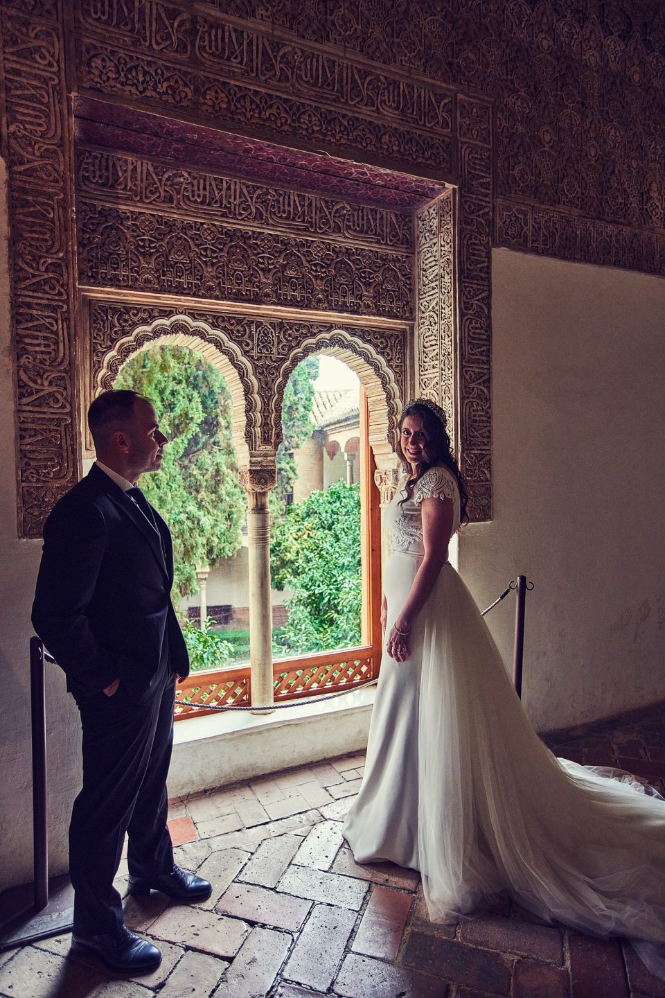 Pareja de novios posando en el interior de un monumento, junto a una ventana con arcos decorados y vistas a un jardín.