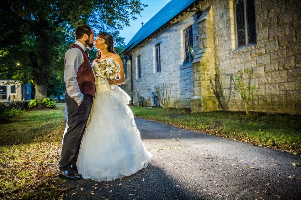 A bridal couple pose and kiss beside a stone building with stained-glass windows.