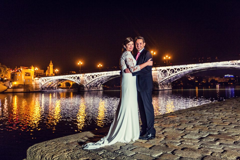 A couple poses at night beside a river, with an illuminated bridge and its reflections in the water in the background.