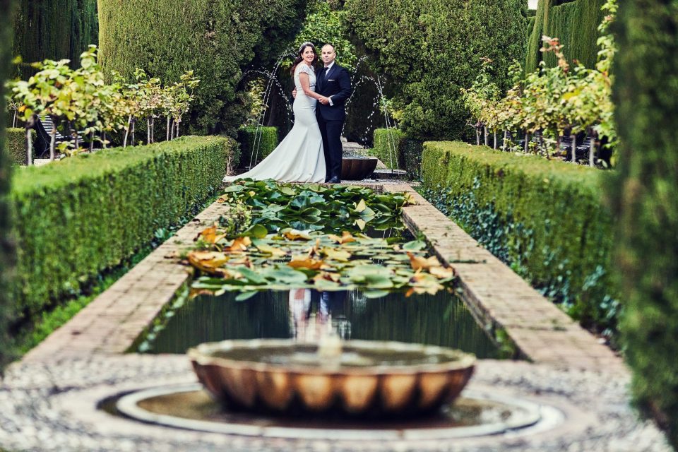 Pareja posando junto a una fuente en un jardín cuidado, rodeada de setos recortados.