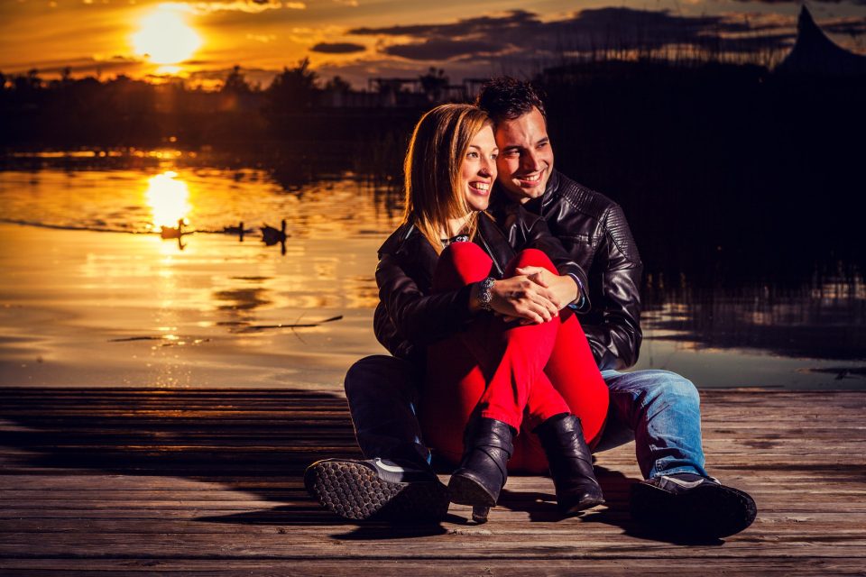 Pareja sentada y abrazada sobre un muelle de madera junto a un lago, sonriendo mientras contempla el horizonte.