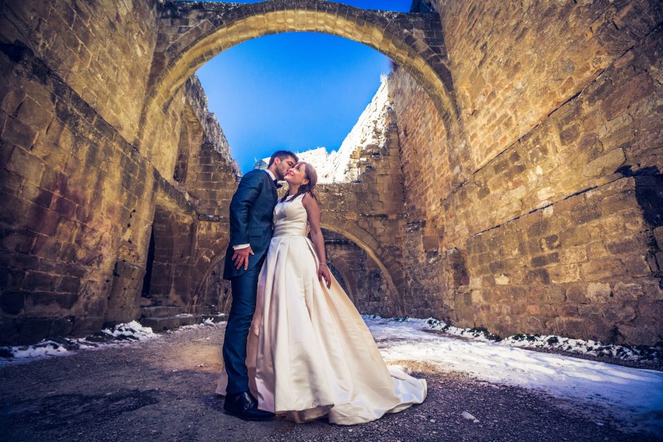 A wedding-dressed couple poses beneath an ancient stone arch.