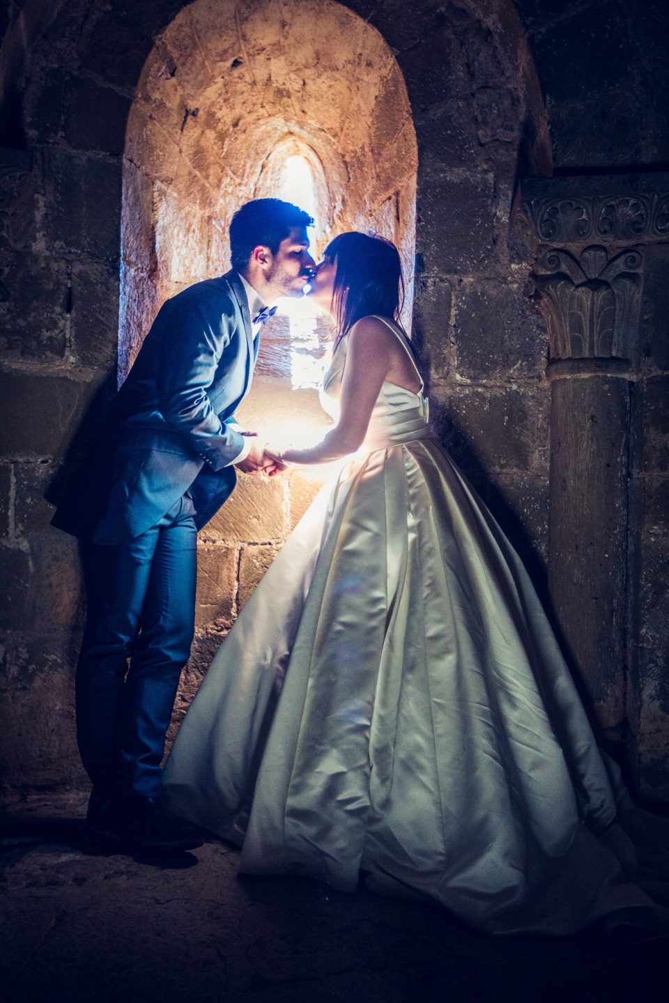 An elegantly dressed couple kiss and hold hands inside a stone building.