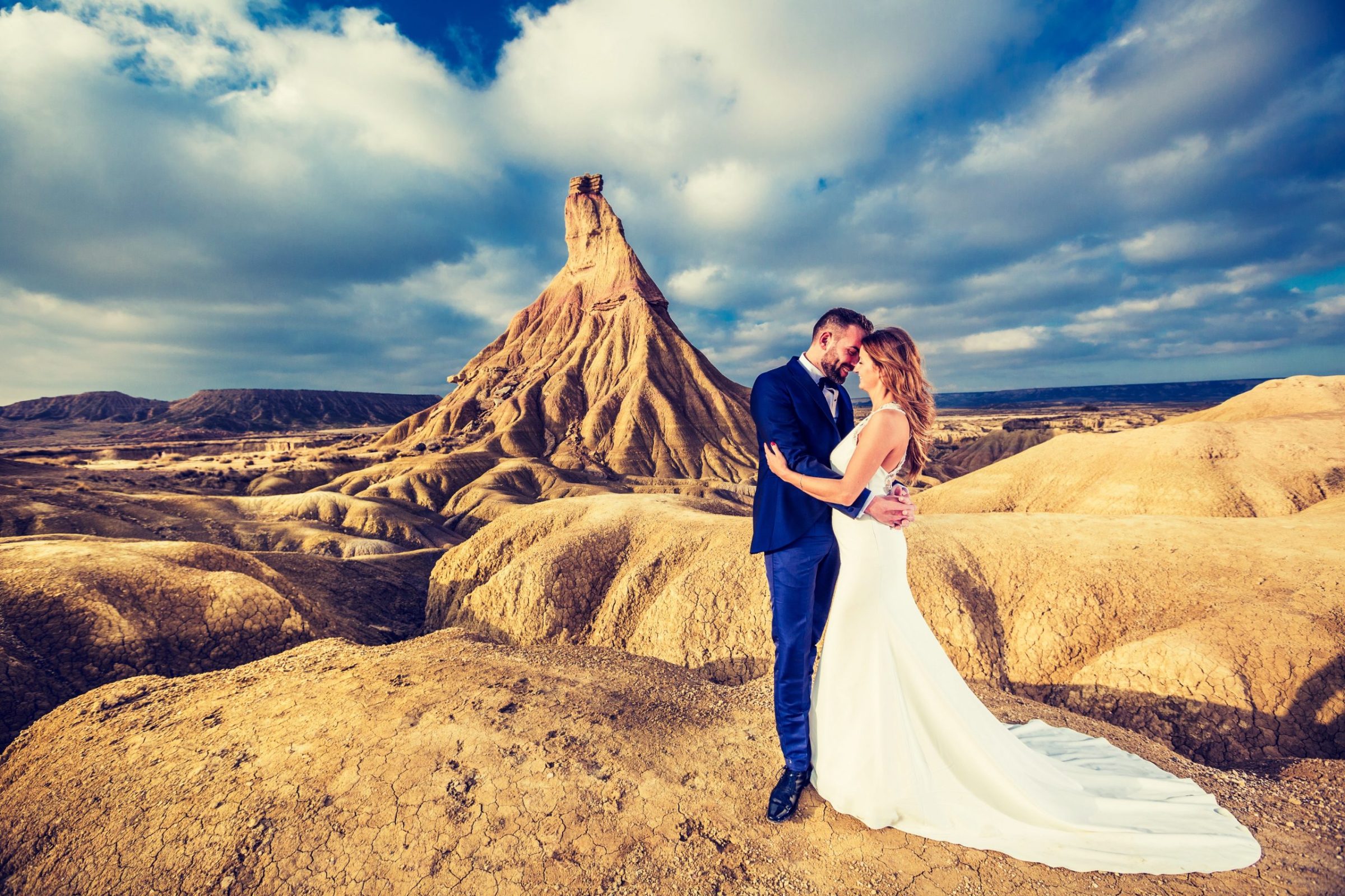 Pareja vestida de novios abrazada en un paisaje exterior árido.