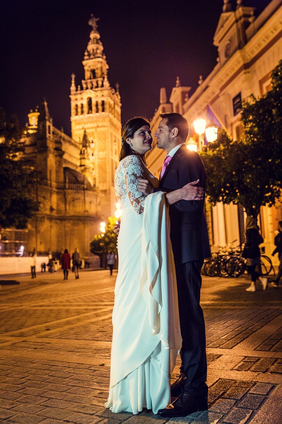 A couple dressed for a wedding pose embracing at night in a paved square.