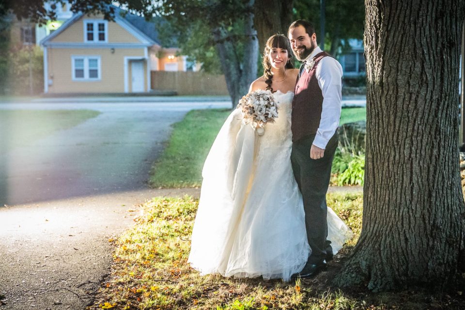 A couple dressed for a wedding poses outdoors beside a large tree.