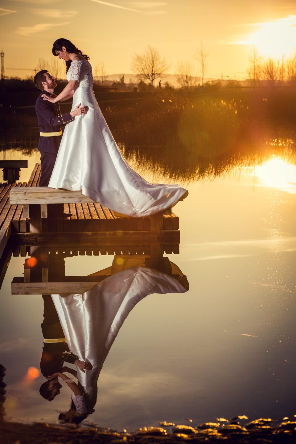 Pareja vestida para una boda sobre un muelle de madera junto a un lago al atardecer.