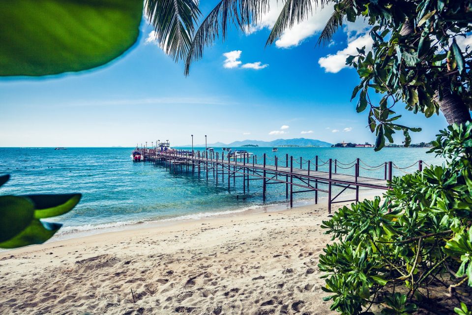 A tropical beach with pale sand and a wooden pier extending into the turquoise sea.