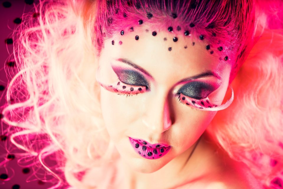 Primer plano de una modelo con los ojos cerrados, maquillaje de fantasía en tonos rosa y negro, sombras brillantes.