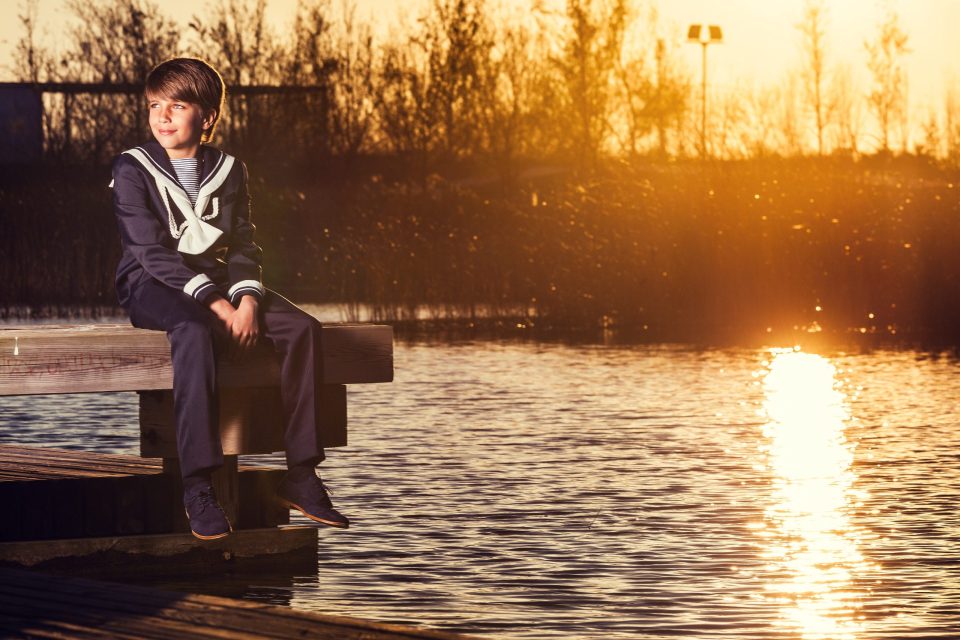 Retrato de un niño sentado en un embarcadero de madera junto al agua, vestido con un traje marinero de comunión.