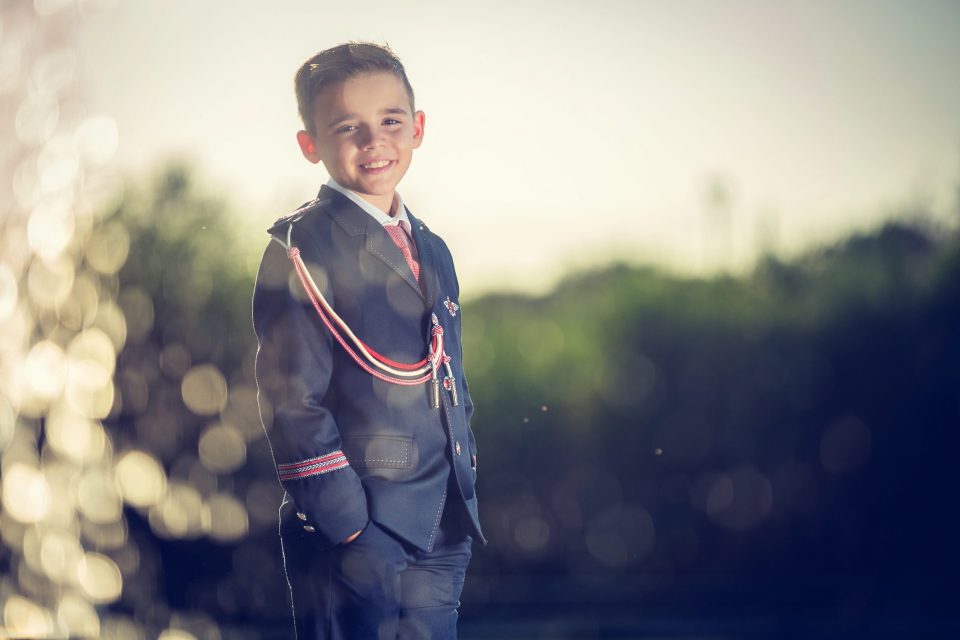 Retrato de un niño sonriente con traje formal azul marino, corbata roja y cordones decorativos.