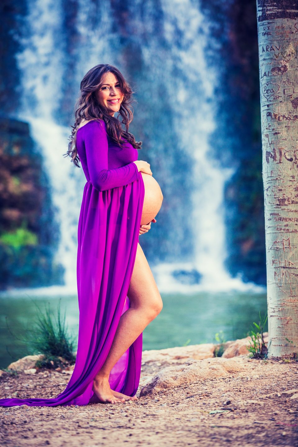 Retrato de una mujer embarazada con un vestido largo morado, de pie junto a un curso de agua con una cascada al fondo.