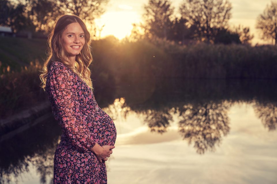 Retrato de una mujer embarazada con vestido de flores, sonriente y con las manos sobre el vientre.