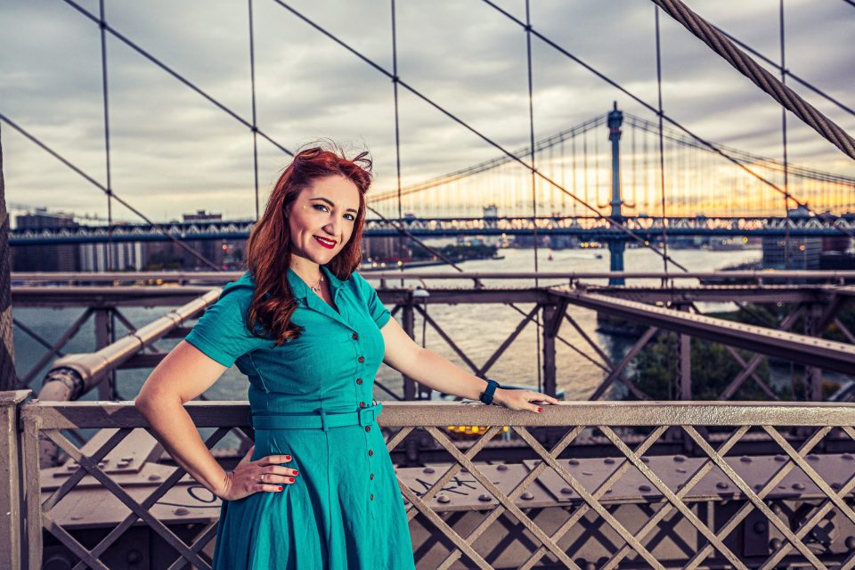 A red-haired woman in a turquoise dress poses beside a bridge railing.