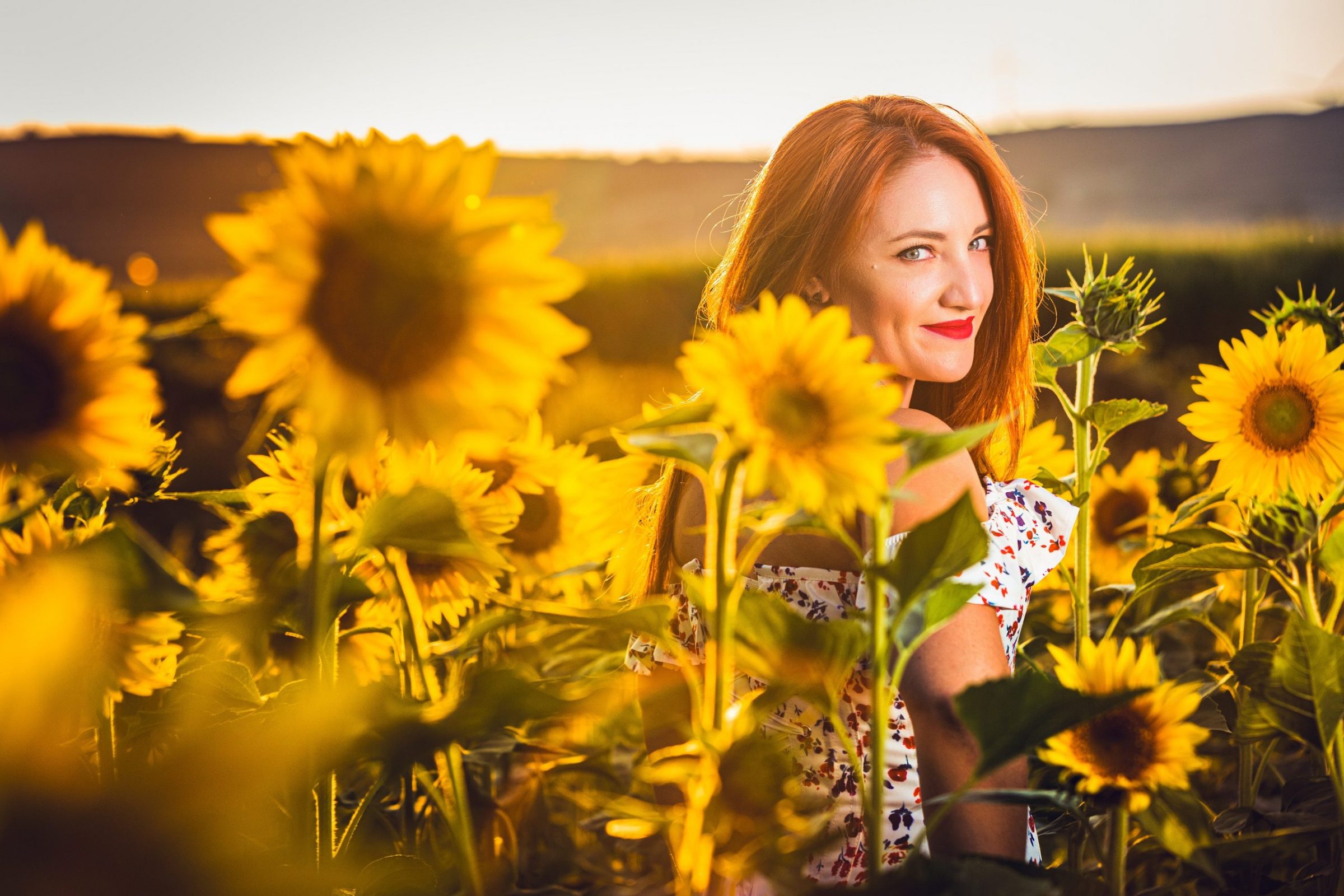 A red-haired woman is pictured among sunflowers in a field bathed in warm evening light.