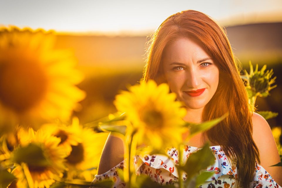 Portrait of a red-haired woman among sunflowers, illuminated by a warm golden light.