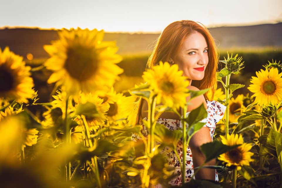 A red-haired woman stands among sunflowers in warm light.